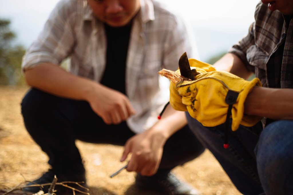 Two adults practicing bushcraft activities outdoors with gloves and knife skills.