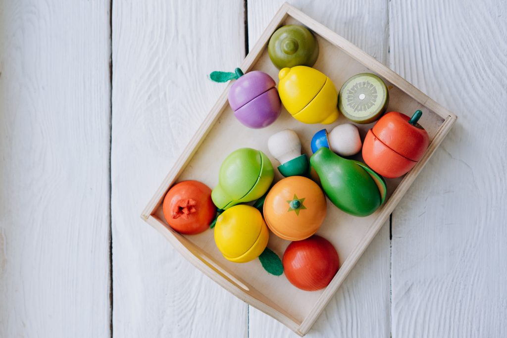 A top view of bright wooden toy fruits and vegetables in a tray on a wooden surface.